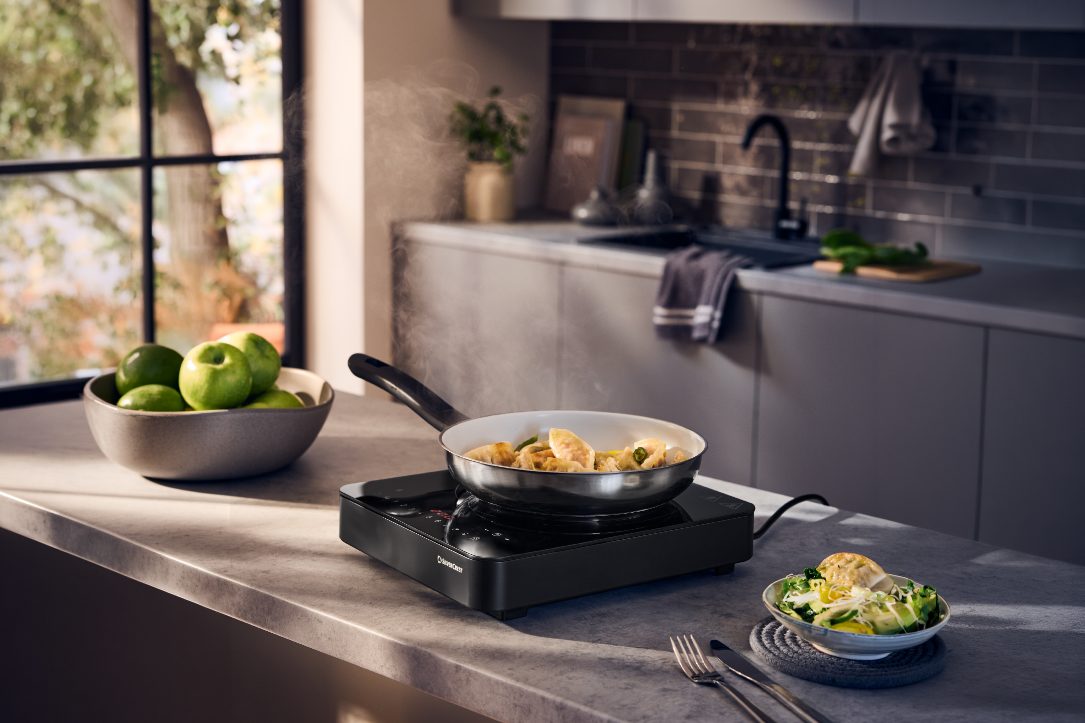 A portable induction hob with a frying pan cooking dumplings, next to a bowl of green apples.