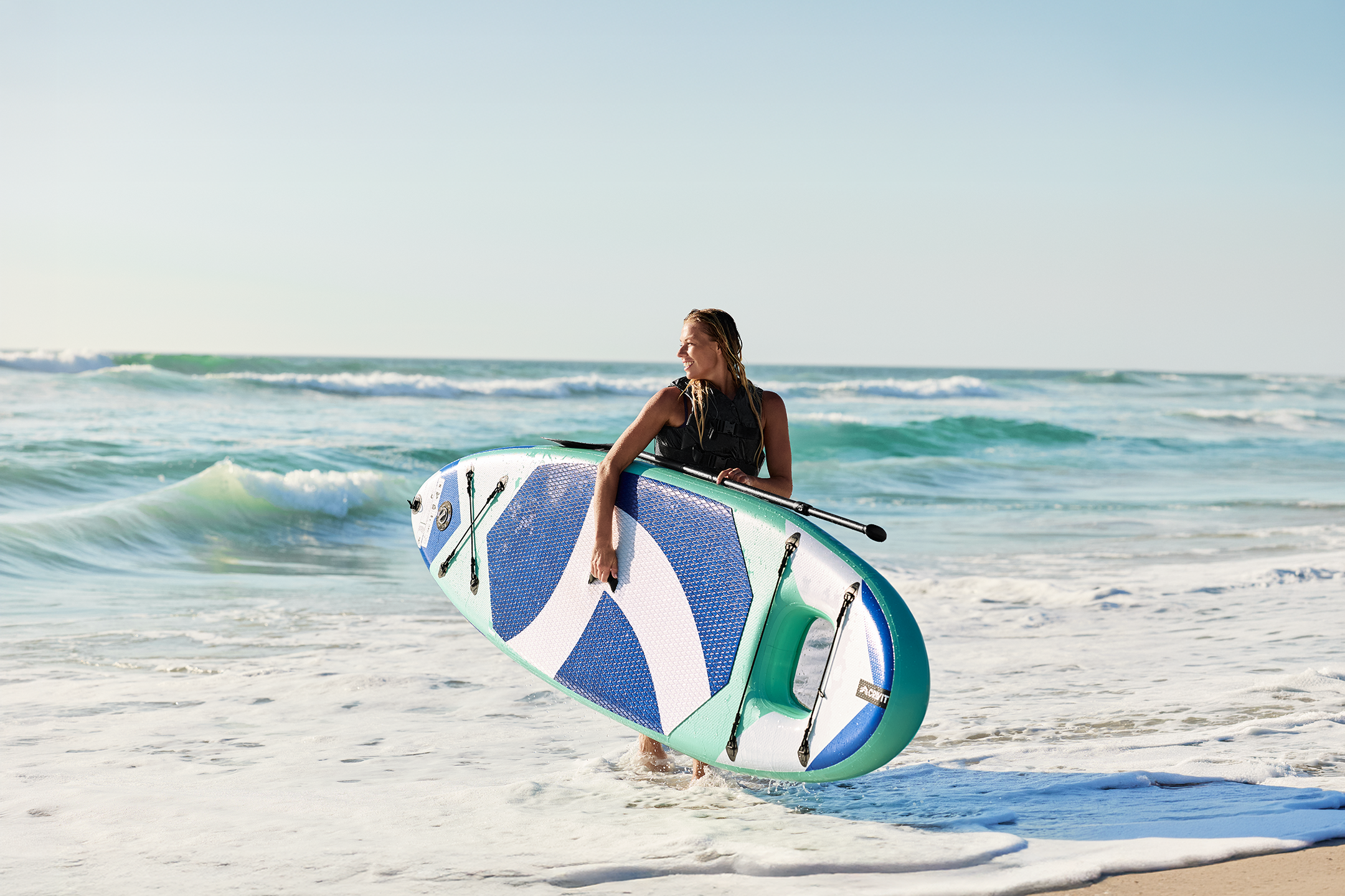 Woman in a life vest holding a paddleboard with a paddle on a beach.