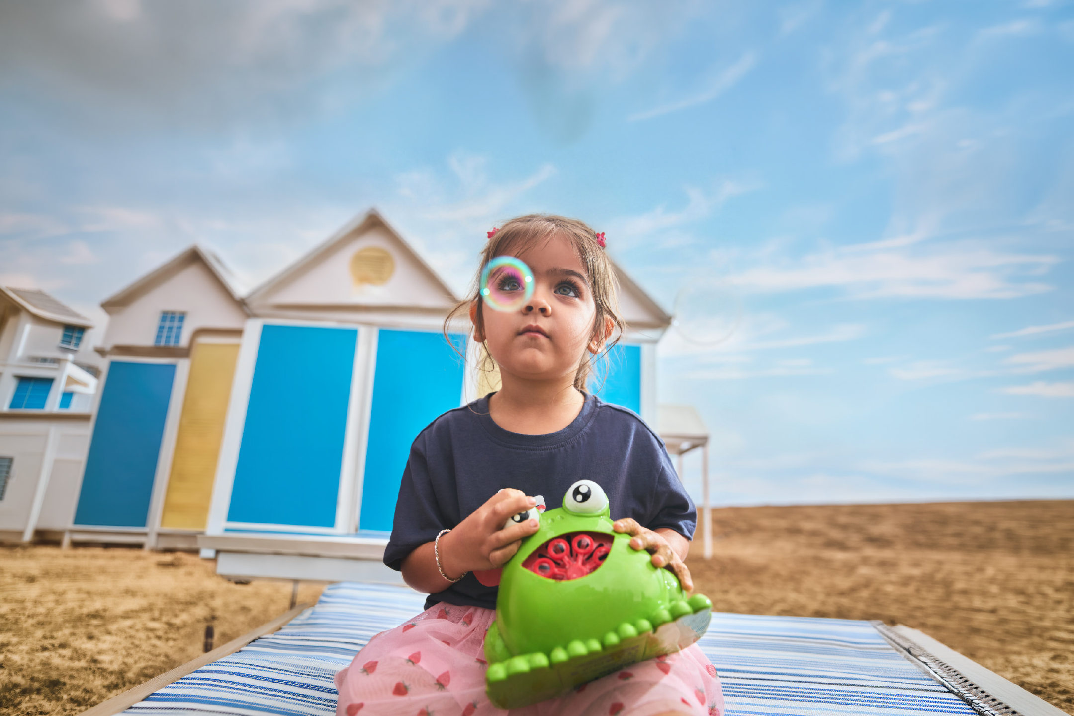 A young girl playing with a green frog-shaped bubble machine on a striped beach mat.