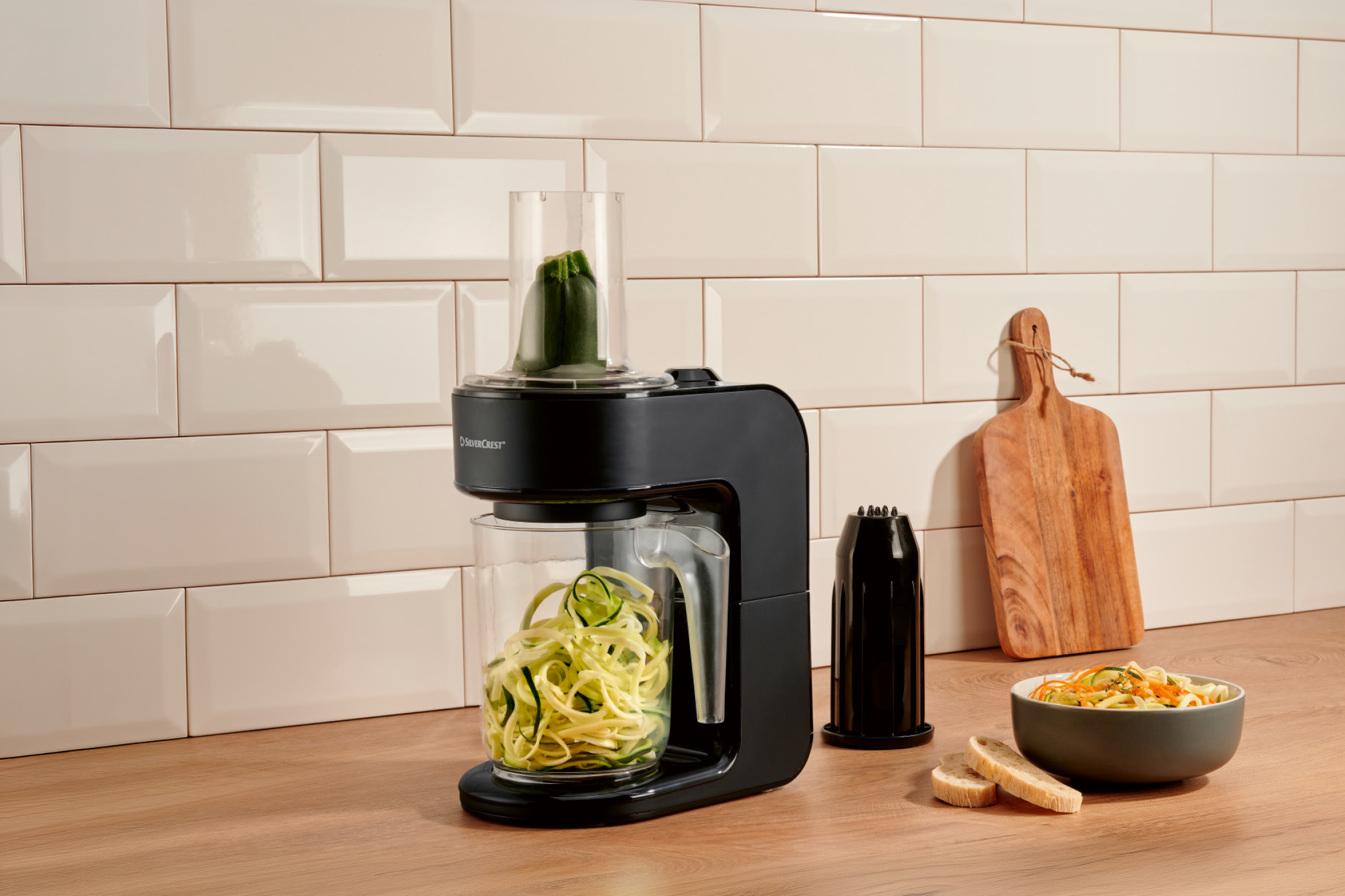 Electric spiralizer making zucchini noodles, with a bowl of vegetable pasta and bread slices.