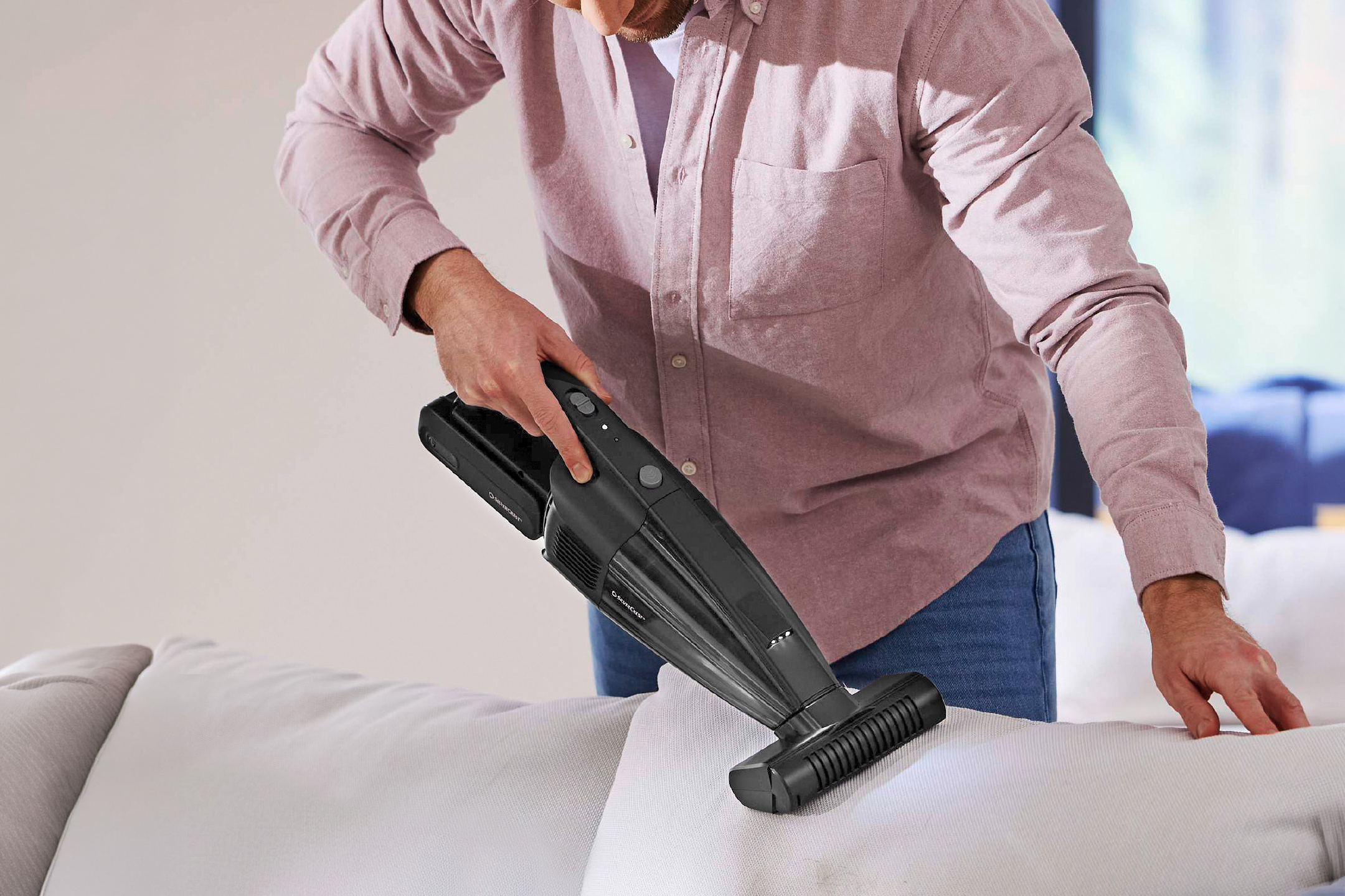 Man cleaning a white sofa with a handheld vacuum cleaner.