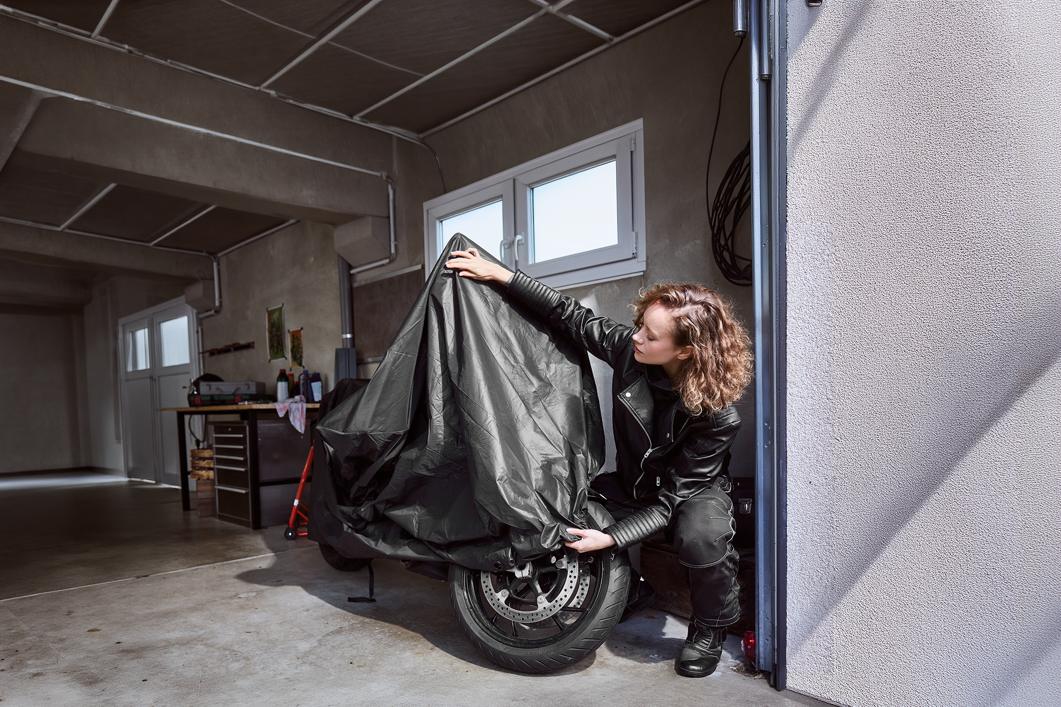 Woman in a leather jacket covering a motorcycle with a protective cover in a garage.