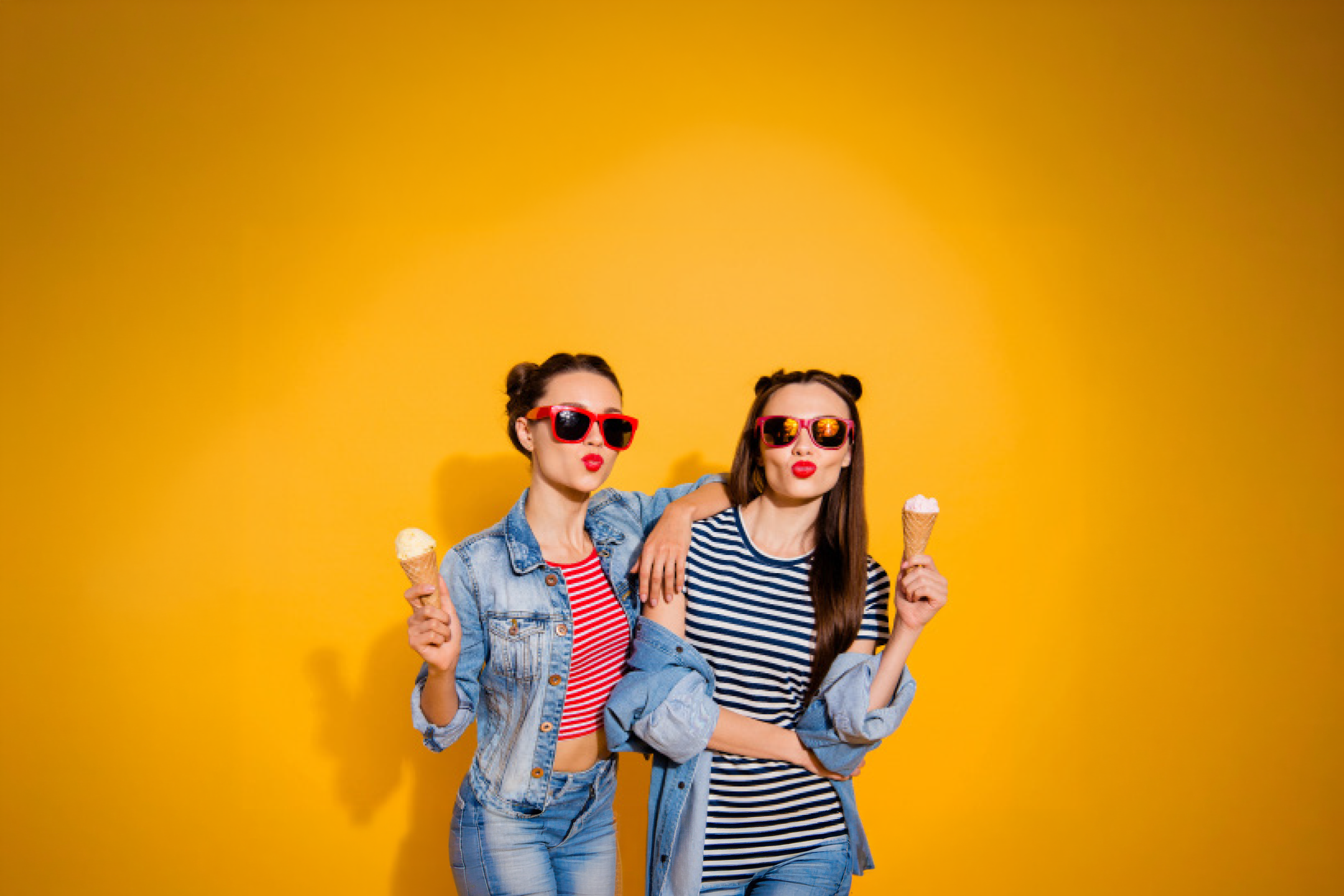 Two young women in sunglasses and denim jackets, holding ice cream cones, making kissy faces.