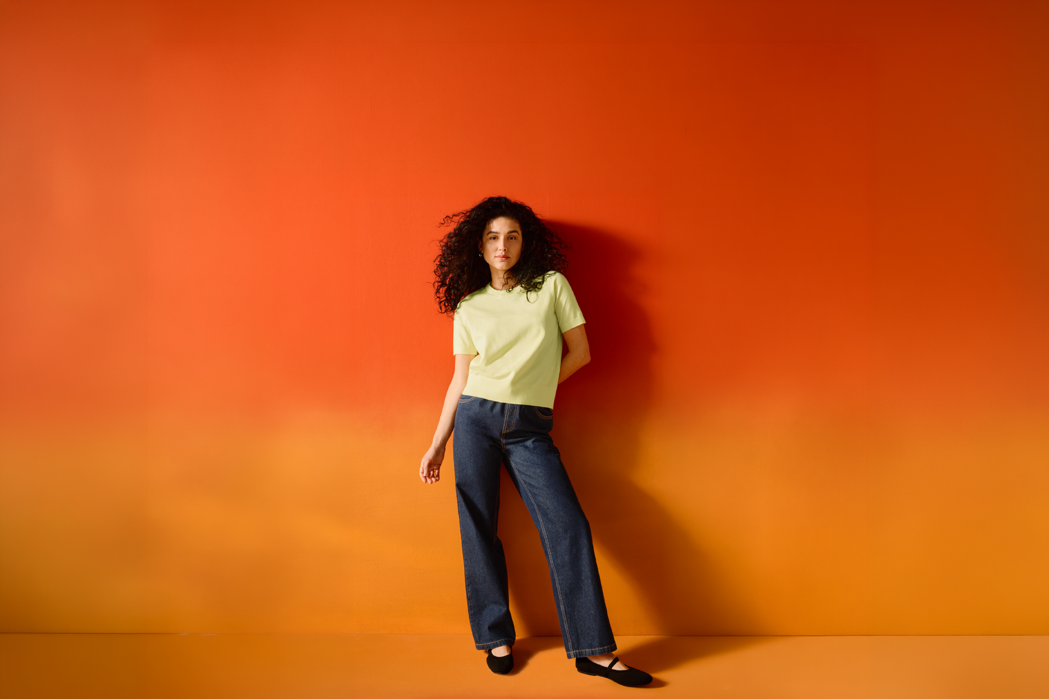 A woman in a light green t-shirt and jeans stands against an orange wall.