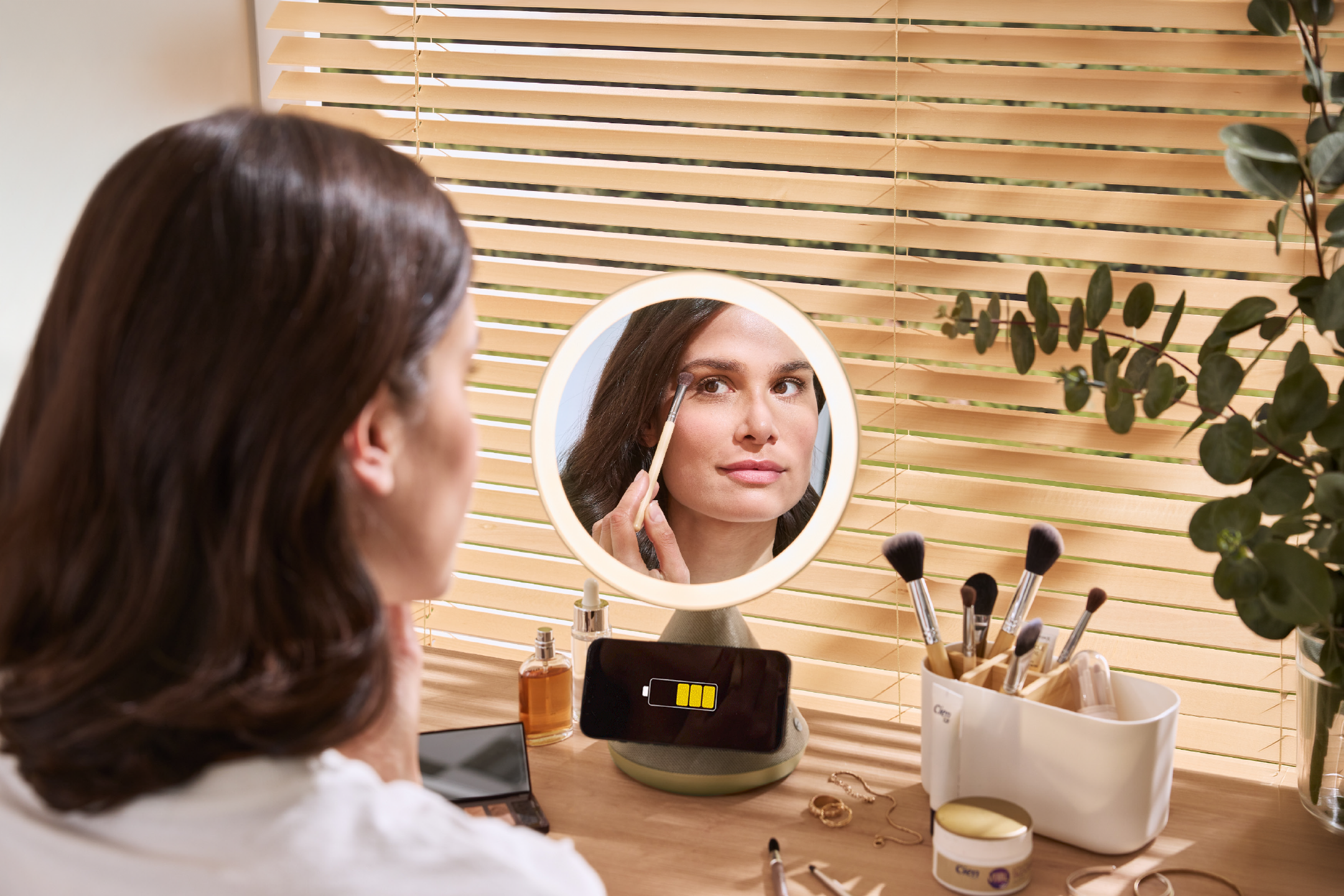 Woman applying makeup in a lighted mirror, with a phone charging on its base.