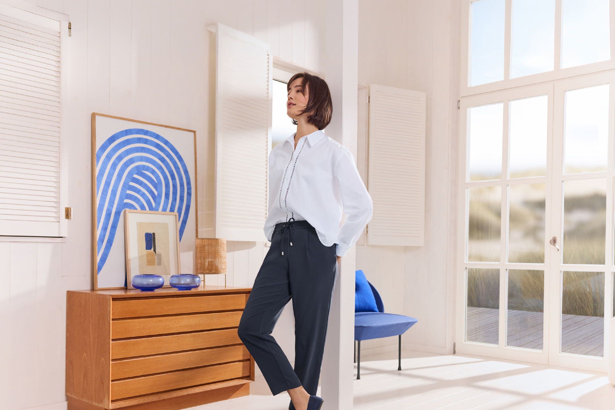 Γυναίκα με λευκό πουκάμισο και σκούρο παντελόνι σε φωτεινό δωμάτιο με ξύλινα έπιπλα και μπλε λεπτομέρειες.