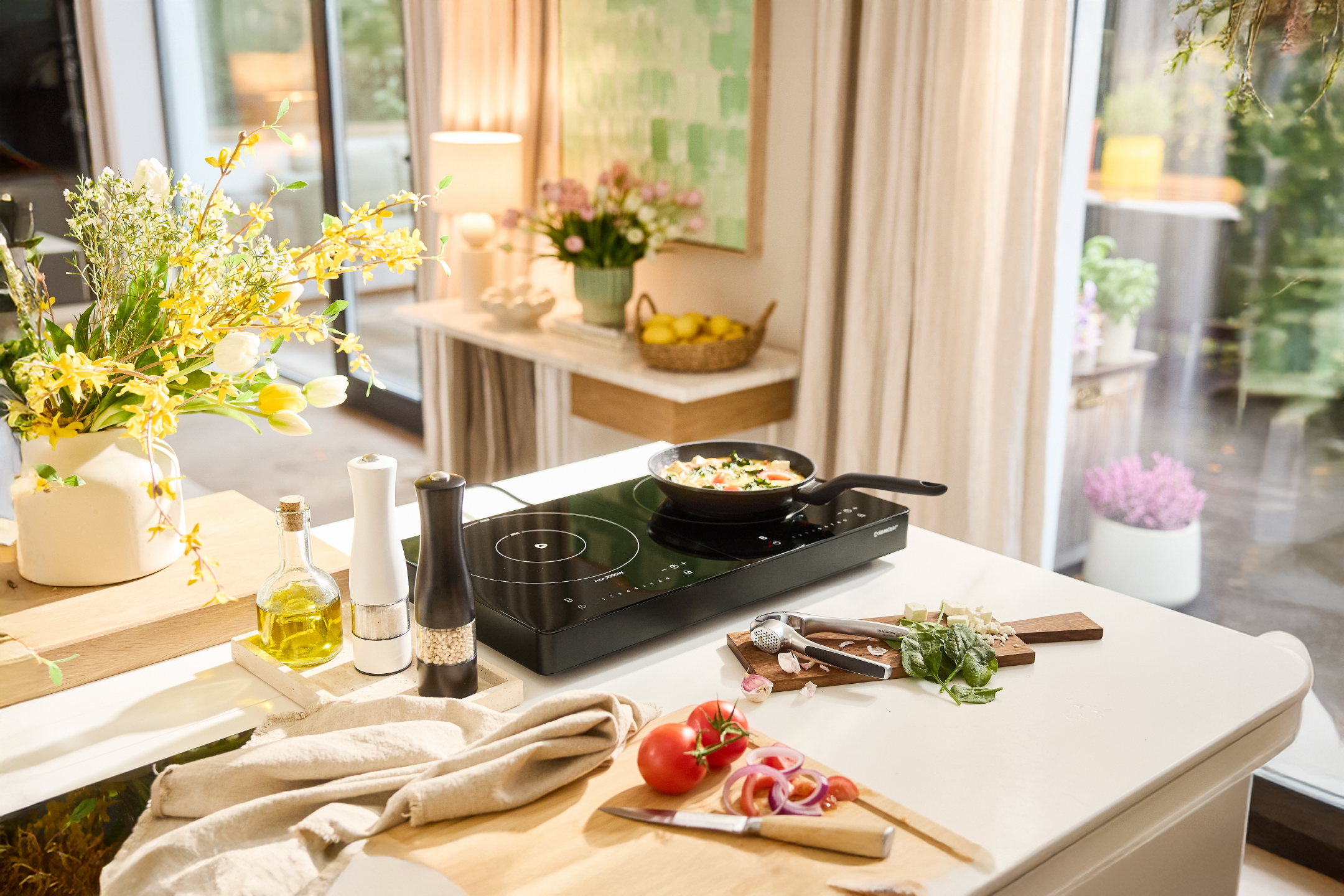 Πάγκος κουζίνας με φορητή επαγωγική εστία, τηγάνι, φρέσκα λαχανικά και λουλούδια.