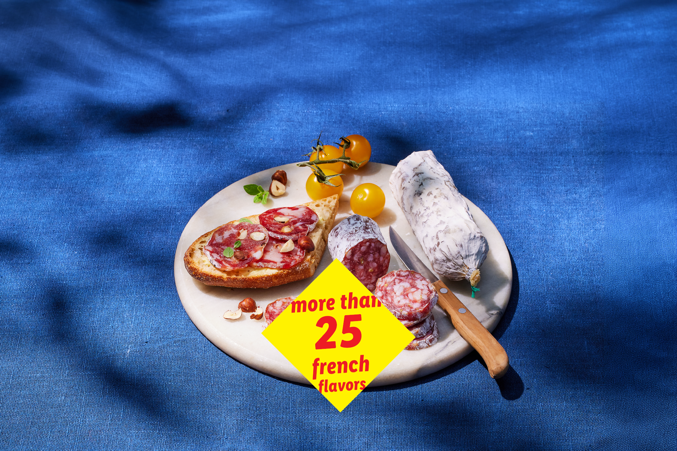 Salami, bread, and tomatoes on a marble board, with text: 'more than 25 french flavors'