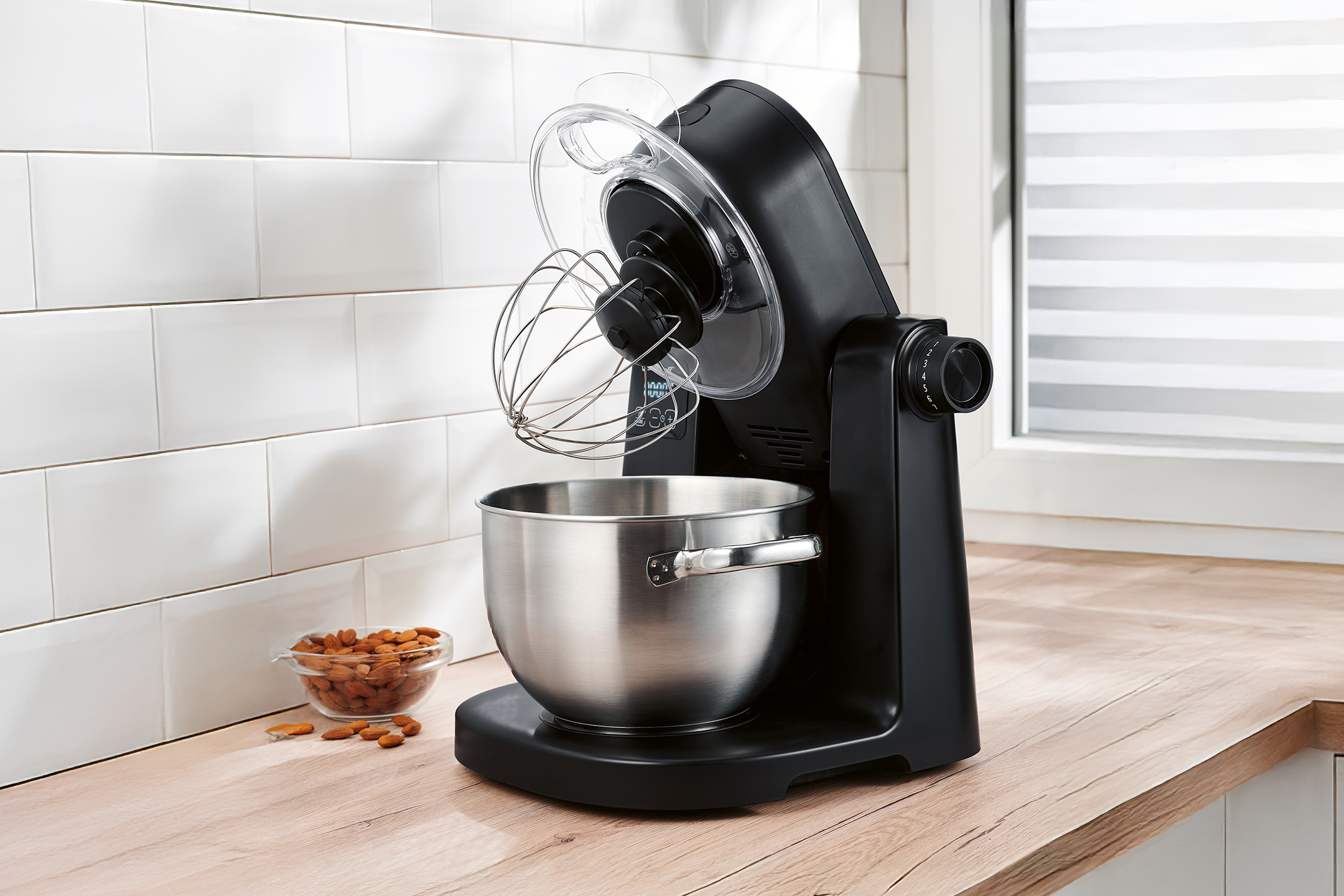Black stand mixer with a stainless steel bowl and whisk, next to a bowl of almonds.