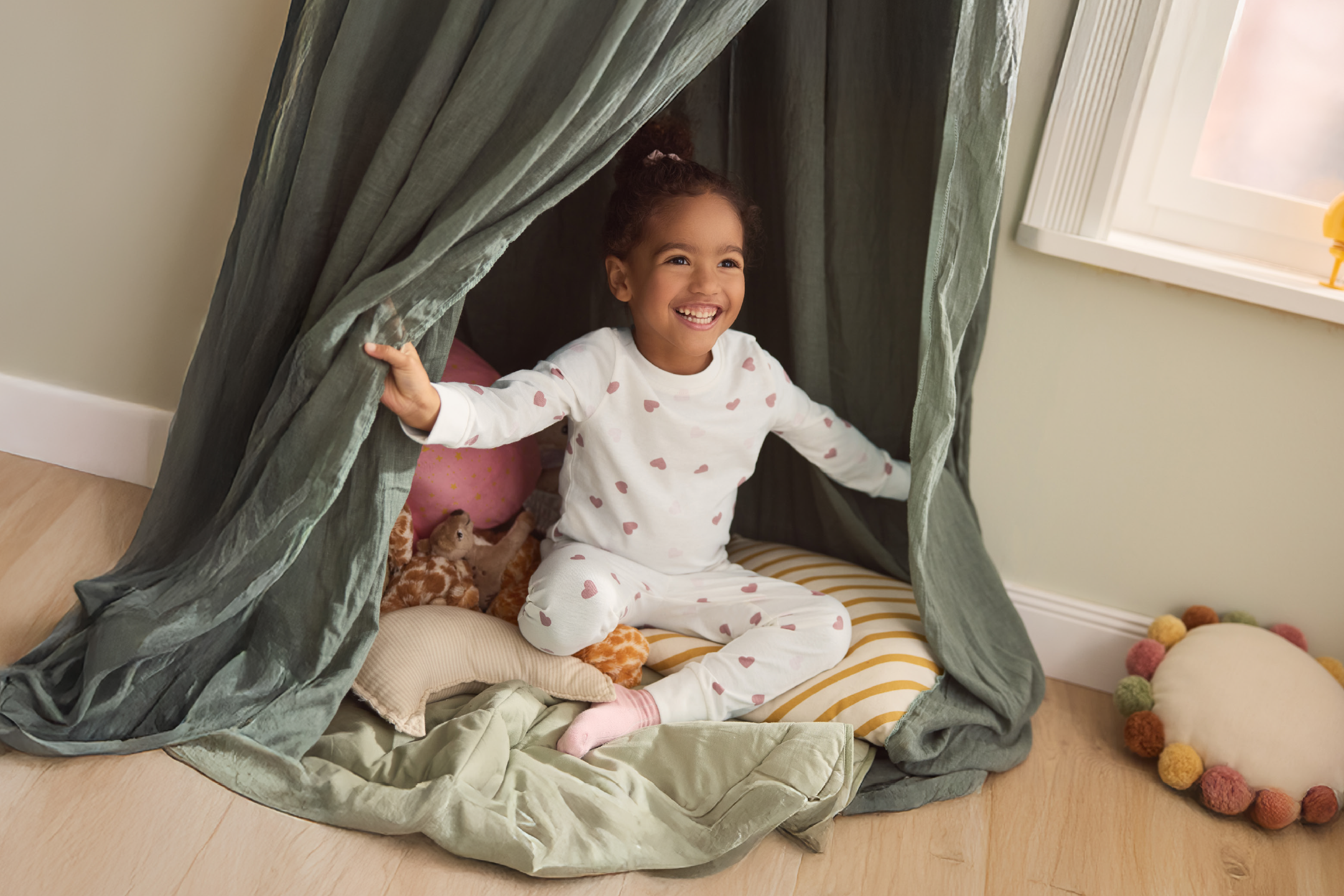 Happy child in heart-patterned pajamas playing in a cozy tent with pillows.