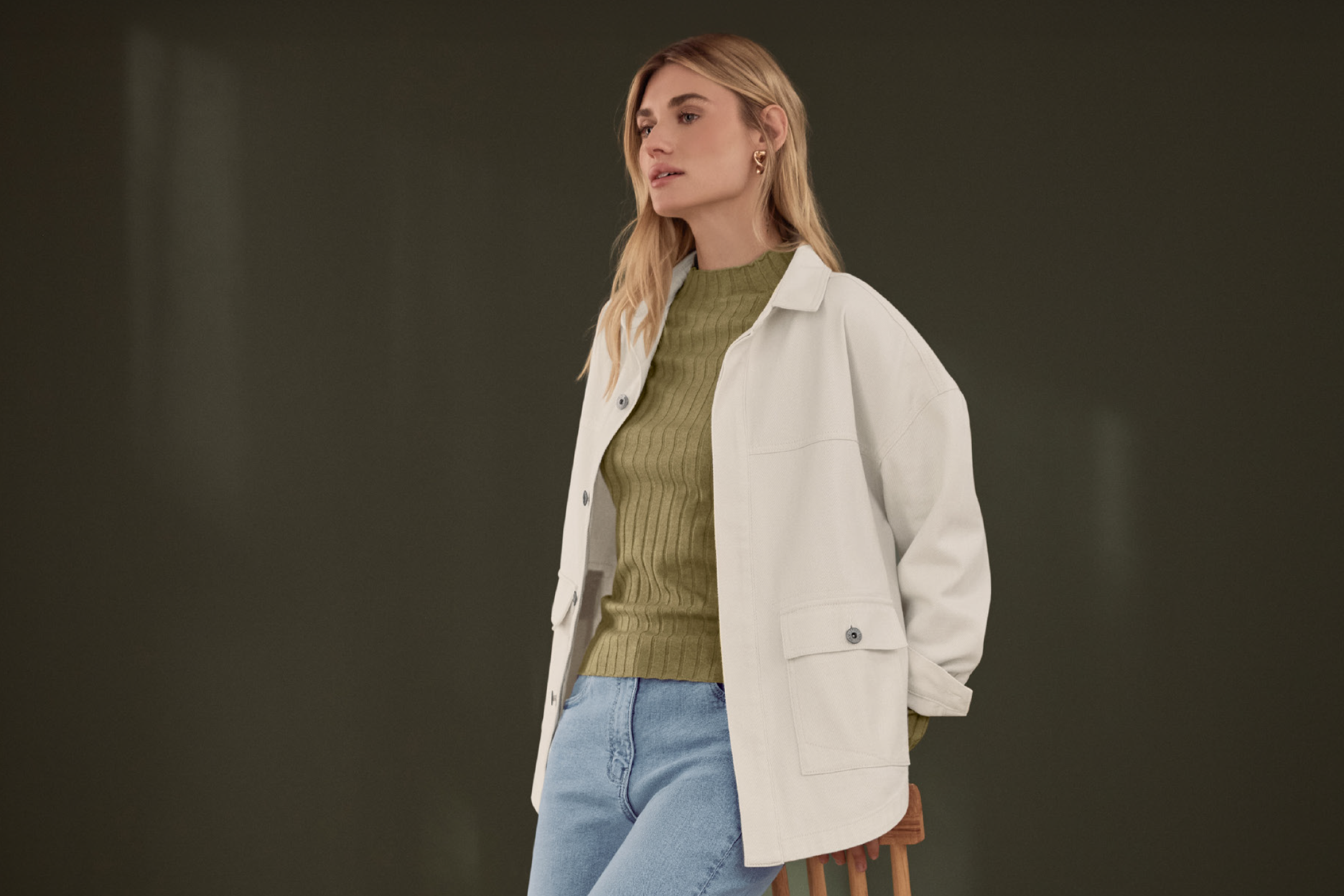 Γυναίκα με λευκό τζάκετ, πράσινο ριμπ τοπ και μπλε τζιν, κάθεται σε ξύλινο σκαμπό.