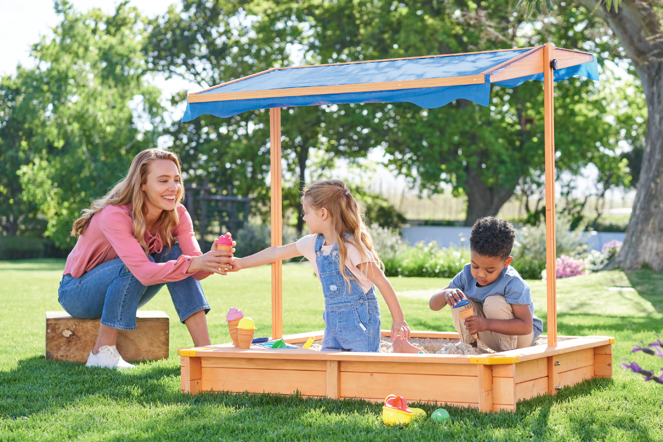 Γυναίκα και παιδιά παίζουν σε ξύλινη αμμοδόχο με μπλε τέντα, φτιάχνοντας παγωτό από άμμο.