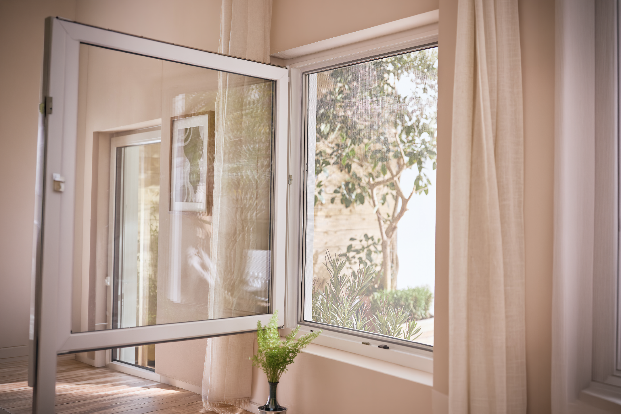 Ανοιχτό παράθυρο με θέα σε πράσινα φυτά και έναν κορνιζαρισμένο πίνακα να αντανακλάται στο τζάμι.