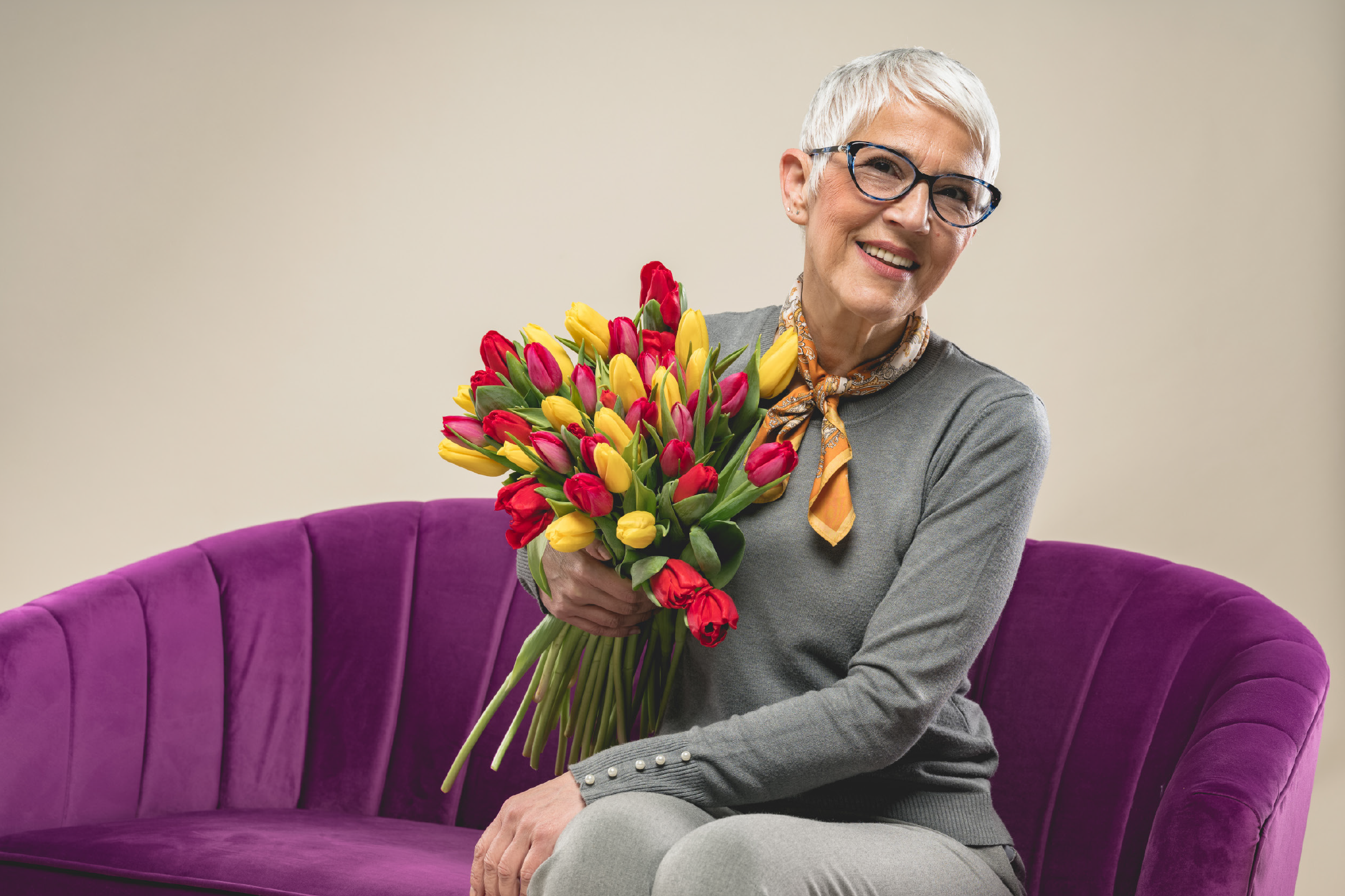Χαμογελαστή ηλικιωμένη γυναίκα με κοντά λευκά μαλλιά και γυαλιά, κρατώντας μια μεγάλη ανθοδέσμη με κόκκινες και κίτρινες τουλίπες, καθισμένη σε έναν μοβ καναπέ.