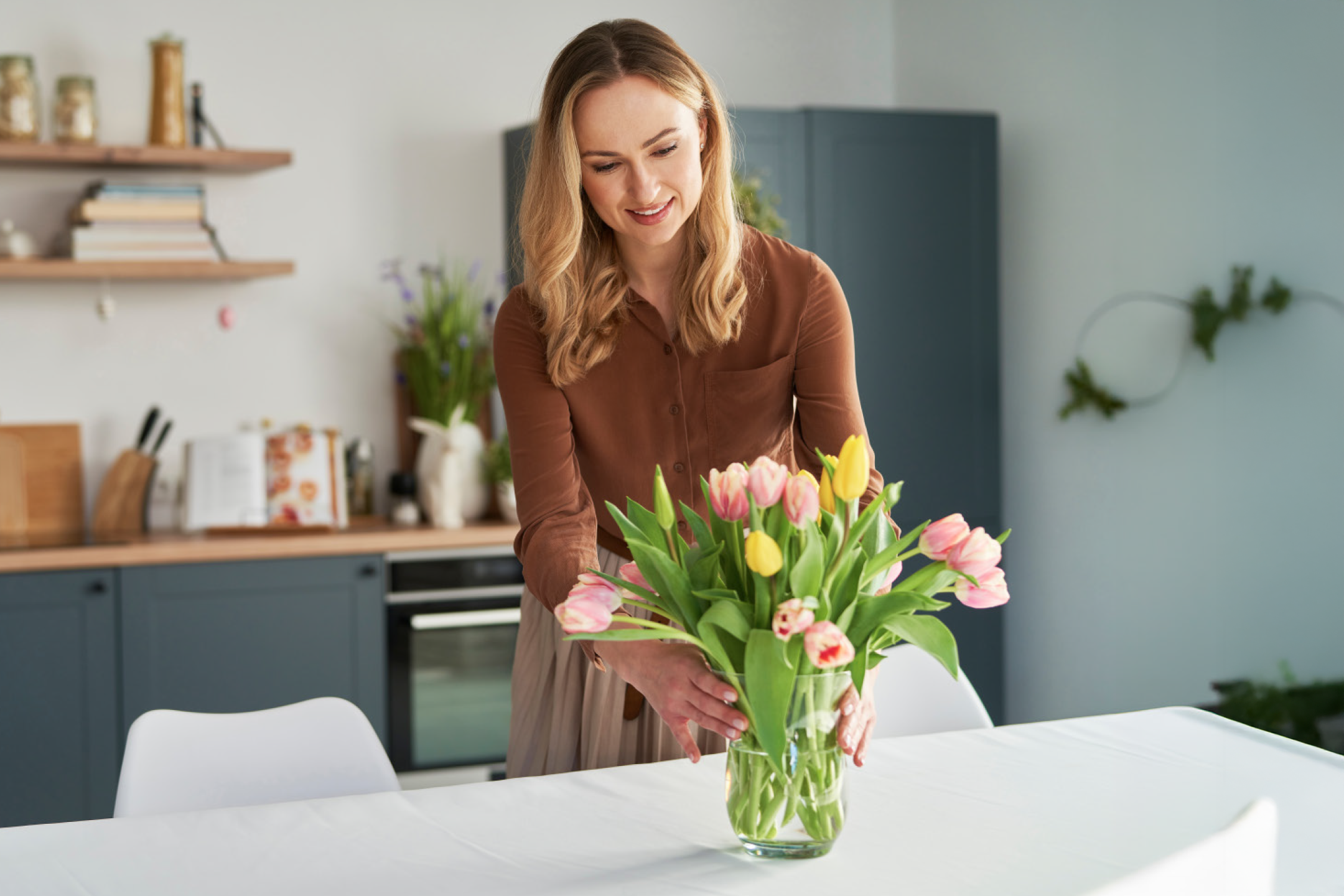 Woman arranging a vase of pink and yellow tulips on a white tablecloth in a kitchen.
