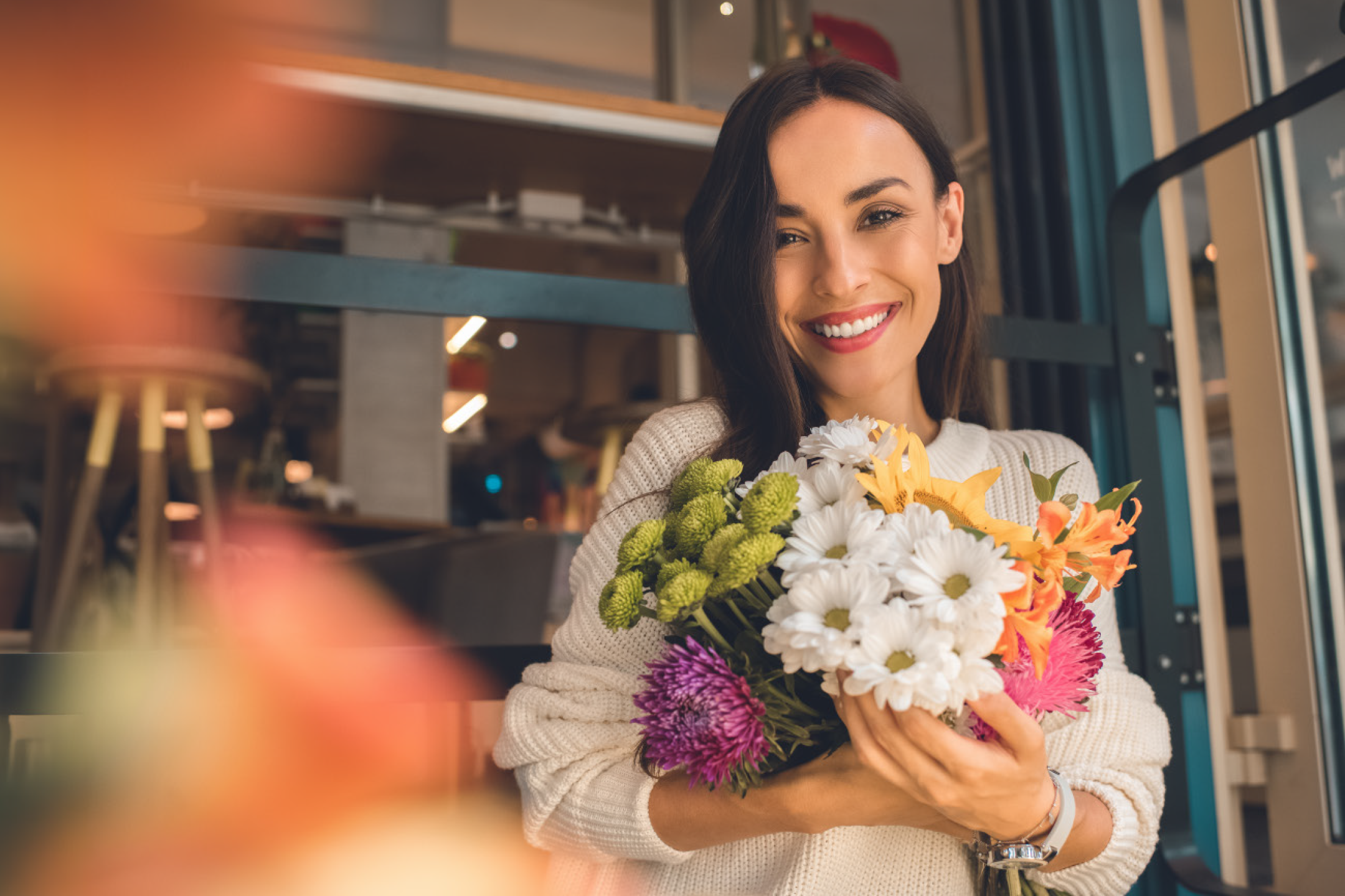 Χαμογελαστή γυναίκα με λευκό πουλόβερ κρατάει μια πολύχρωμη ανθοδέσμη.