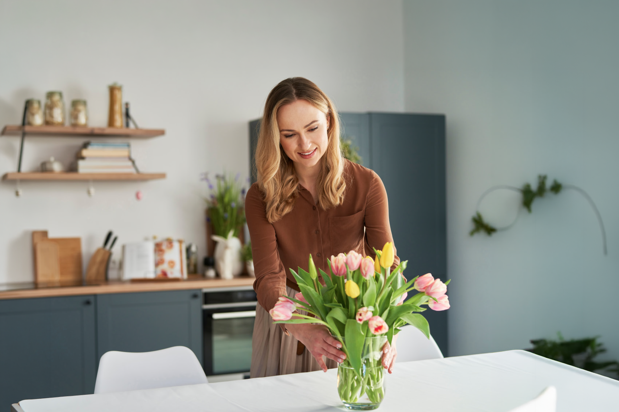 Woman arranging a vase of fresh tulips on a white table in a modern kitchen.