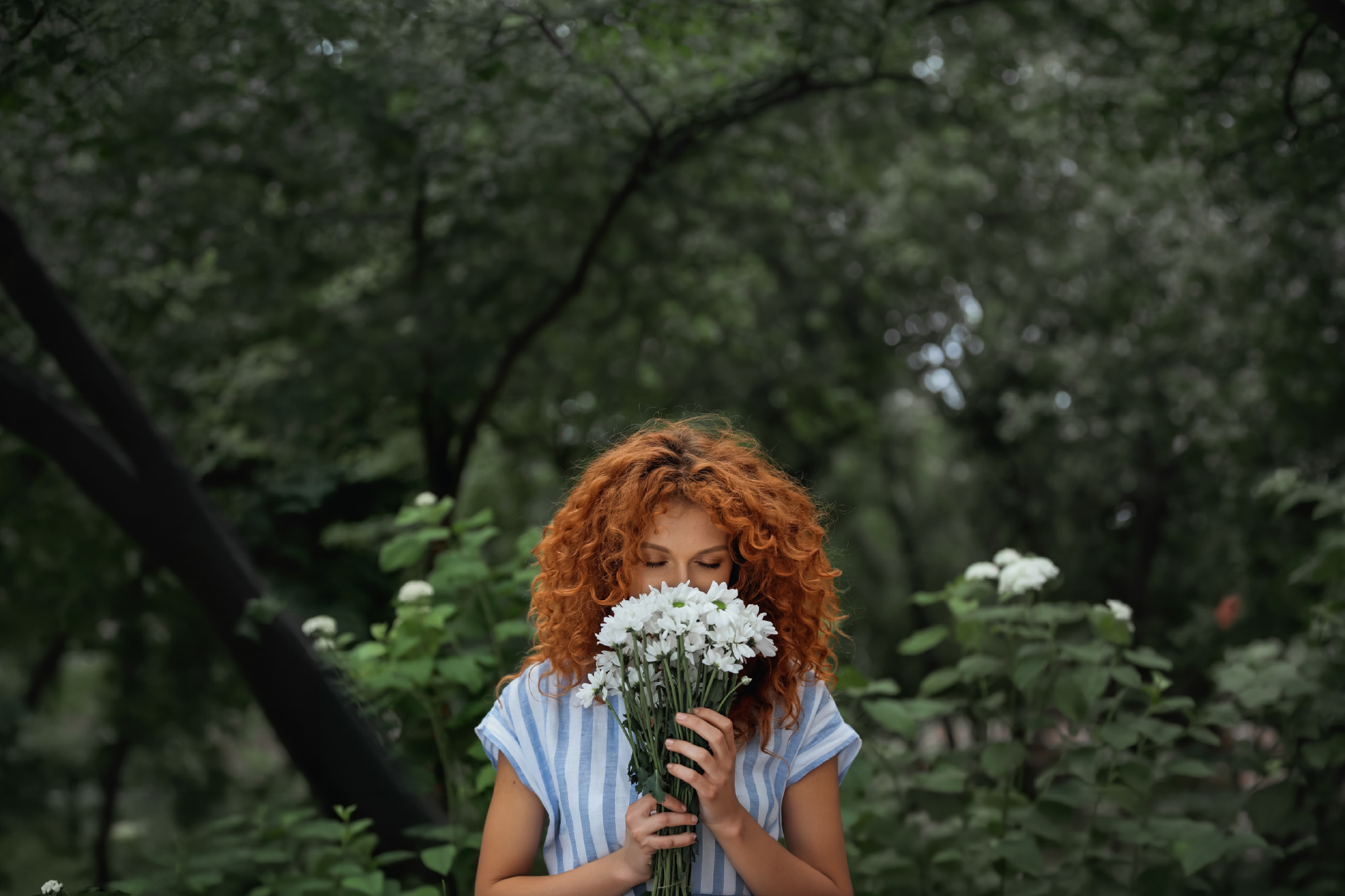 A woman with red curly hair smelling a bouquet of white flowers in a garden.