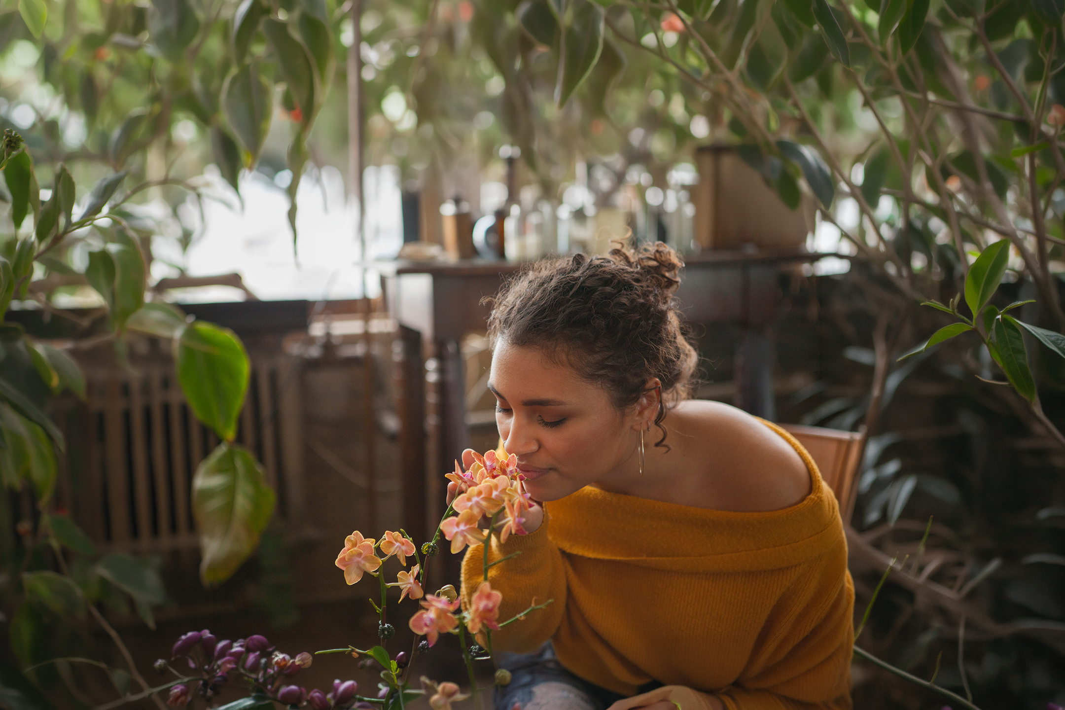 Woman in a yellow sweater smelling flowers in a greenhouse.