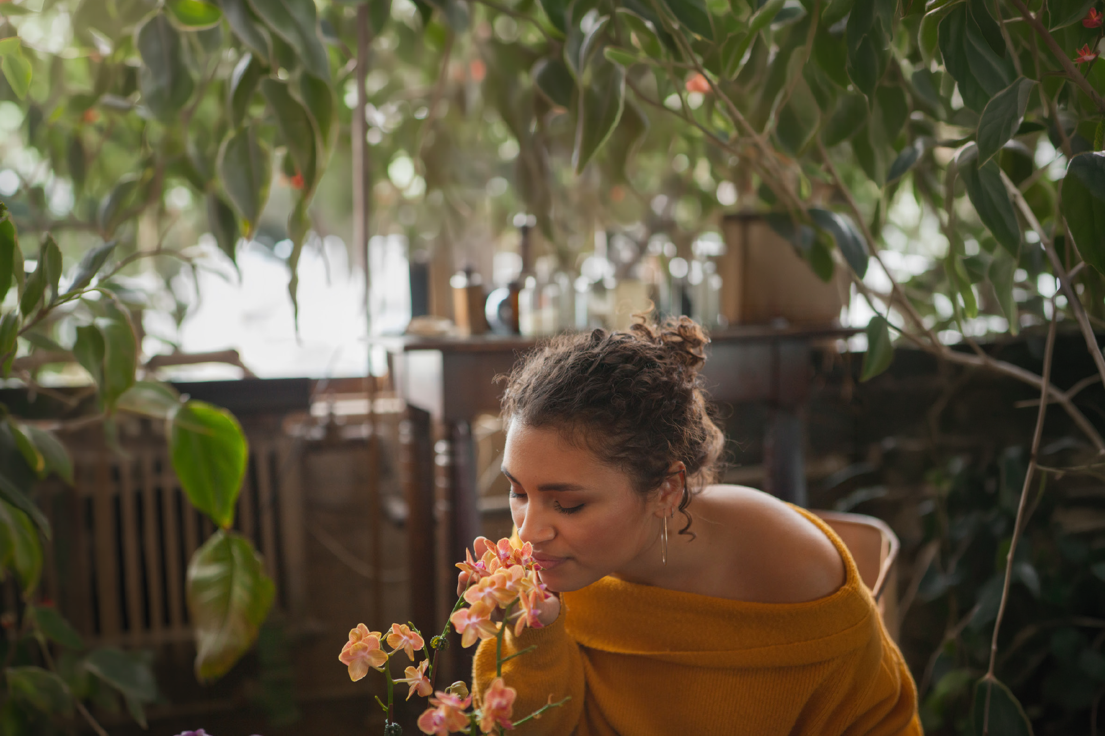 A woman smelling orange orchids in a cozy greenhouse with lush green plants.