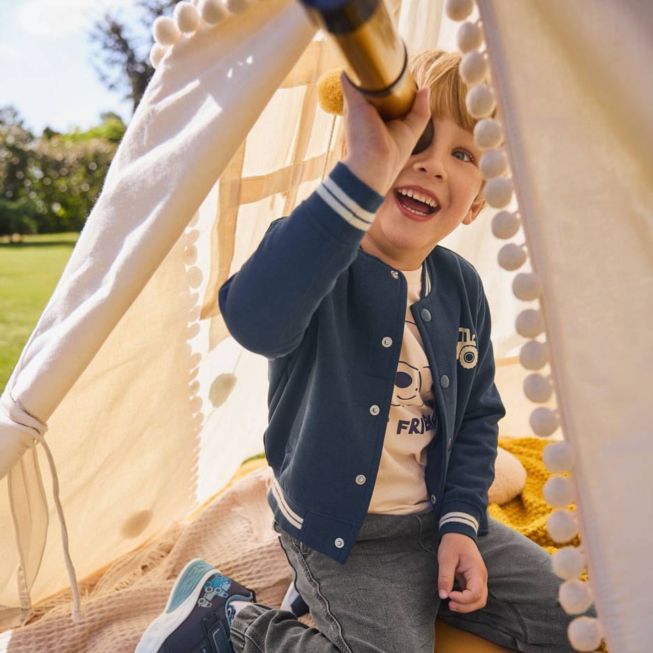 Smiling boy in blue jacket and beige t-shirt, looking through a telescope in a tent.