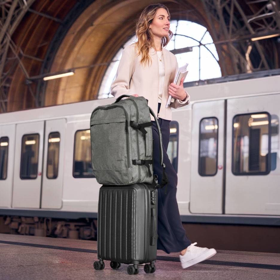 Woman with a grey backpack and black wheeled suitcase on a subway platform.