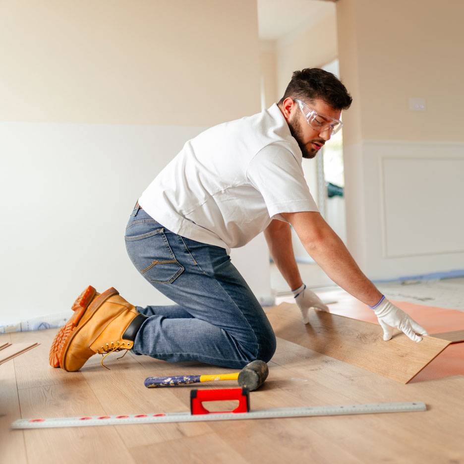 Άνδρας τοποθετεί laminate δάπεδο, φορώντας γυαλιά ασφαλείας και μπότες εργασίας.