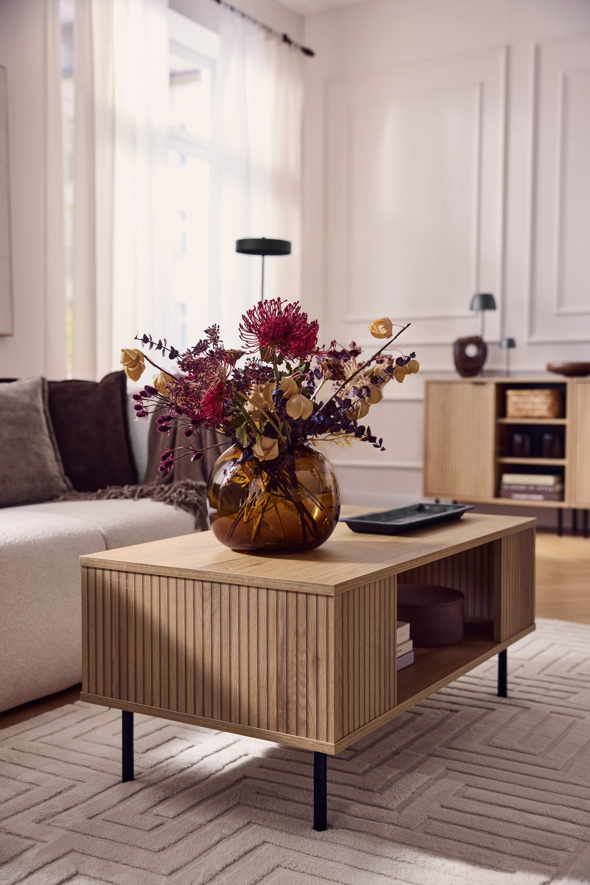 Wooden coffee table with a fluted front, a vase with dried flowers, and a beige sofa.